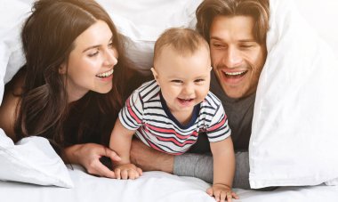 Mutlu bir aile yatakta beyaz bir battaniyenin altında peekaboo oynuyor. Ebeveynler gülümsüyor ve bebeklerine bakıyor. O da gülüyor ve kameraya uzanıyor..