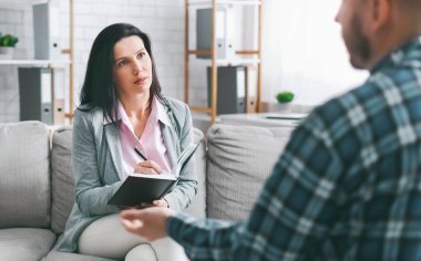 Gri ceketli ve beyaz bluzlu bir kadın ofisteki bir koltukta oturup terapi seansı sırasında notlar alıyor. Terapist onun karşısında oturuyor ve odaklanamıyor..