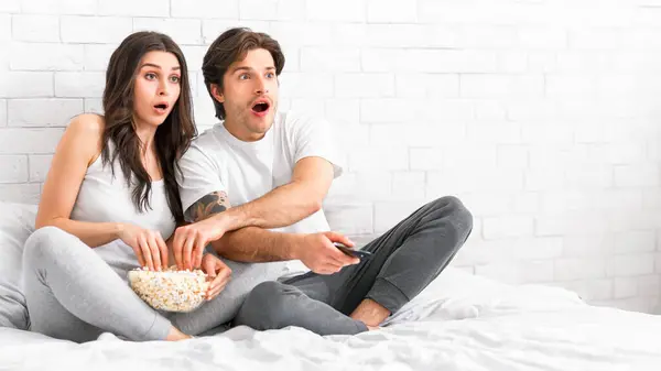 stock image A young couple is sitting on a bed, their faces showing surprise as they watch something on TV. The man holds the remote in his hand, while the woman reaches for popcorn in a bowl, copy space