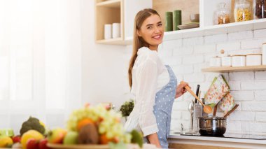 Mavi benekli önlük giyen bir kadın mutfaktaki ahşap kaşıkla ocakta tencereyi karıştırıyor, gülümsüyor ve omzunun üstünden bakıyor..