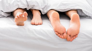İki çıplak ayak görüntüsü, bir erkek ve bir kadın, beyaz yatağın altından sarkıyor, bir çiftin yatakta uyuduğunu gösteriyor..