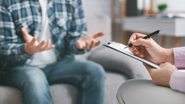 A therapist is seated across from their patient and taking notes on a clipboard with a pen, while the patient gestures with their hands and speaks, cropped
