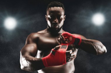 Ciddi bir ifadeye sahip kaslı bir boksör ellerini ışıksız bir boks ringinde kırmızı bant ile sarıyor..