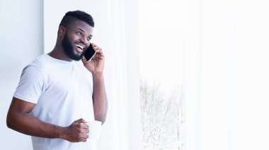 Sakallı Afrikalı Amerikalı adam telefonda konuşurken gülümsüyor. O, elinde bir fincan tutuyor ve bir pencerenin önünde duruyor. Adam beyaz bir tişört giyiyor, fotokopi odası.