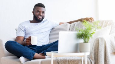 Beyaz tişörtlü ve kot pantolonlu genç bir adam oturma odasındaki koltukta dinleniyor, elinde beyaz bir fincan kahve tutuyor ve pencereden dışarı bakıyor.