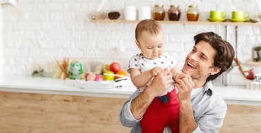 Genç bir baba bebeğini mutfakta tutuyor. İkisi de gülümsüyor ve birbirlerine eşlik etmekten zevk alıyor gibi görünüyorlar. Arkalarında meyveler ve diğer eşyalar var..