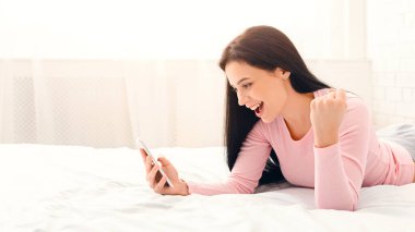 Genç bir kadın beyaz bir yatak örtüsünün üzerinde karnının üzerinde uzanmış akıllı telefonuna keyifli bir ifadeyle bakıyor. Parlak güneş ışığı odayı dolduruyor. Sabah ya da öğleden sonra olabilir.