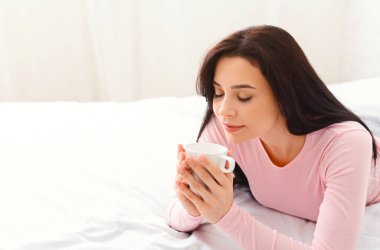 Bir kadın elinde bir fincan kahveyle bir yatakta uzanıyor. Rahatlamış ve rahat görünüyor, sabah içkisinin tadını çıkarıyor..