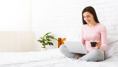 Bir kadın bir yatakta oturmuş elinde bir fincan kahve tutarken dizüstü bilgisayarla çalışıyor. İşine odaklanmış ve meşgul görünüyor, onu çevreleyen rahat ve rahat bir atmosferi var, boş bir alan.