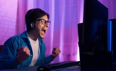 Kulaklık ve gözlük takan genç bir adam bilgisayar ekranının önünde oturmuş video oyunları oynuyor. Heyecanlı ve kollarını kaldırmış zafer kazanmış gibi..