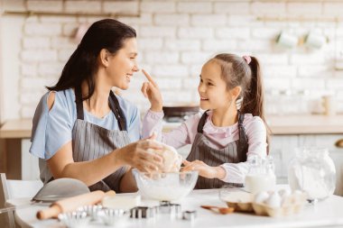 Bir anne ve küçük kızı mutfakta birlikte yemek pişiriyorlar. Anne bir kâsede hamuru karıştırırken kız annesinin burnuna neşeyle dokunuyor. İkisi de önlük takıyor.