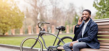 Afrikalı Amerikalı bir işadamı, parktaki bir bankta otururken ve bisikleti yakınlardayken telefonuyla konuşuyor. Ortam sakin ve profesyonel, yeşil, huzurlu bir parkta geçiyor..
