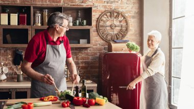 Önlük giyen yaşlı bir çift, beyaz dolaplar ve tuğla aksanlı modern bir mutfakta birlikte yemek pişiriyor. İkisi de gülümsüyor ve birlikte vakit geçiriyorlar..