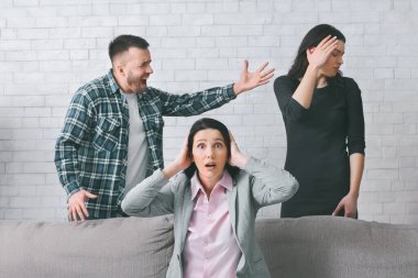 Bir kadın ellerini kulaklarının üzerine koyarak koltukta oturur ve bir erkek kanepenin arkasında duran başka bir kadına bağırır. Kanepenin arkasında eli alnında duran kadın..