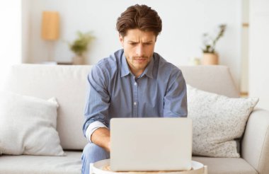 Bir adam kanepede oturur ve dizüstü bilgisayarıyla çalışır. O, işine odaklanmış ve ciddi..