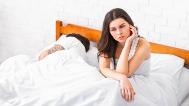 Genç bir kadın yatakta oturuyor, düşünceli ve üzgün görünüyor. Partneri onun yanında mışıl mışıl uyurken, o başını elinin üzerine koyar. Görüntü bir yalnızlık hissi veriyor.