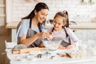 Bir anne ve kızı mutfakta birlikte çalışıyorlar, kurabiye pişiriyorlar. Annesi elinde bir yumurta tutuyor ve onu bir kase un haline getirmek üzereyken kızı gülümseyerek izliyor..