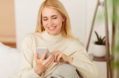 Gülümseyen sarışın bir kadın akıllı telefonunu kapalı bir koltukta otururken kullanır..