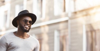 Şapkalı ve güneş gözlüklü bir adam şehir caddesinde yürüyor. Gülümsüyor ve yana bakıyor..