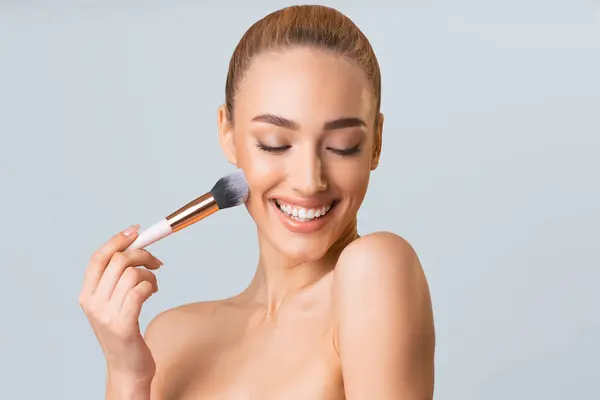 stock image A young woman with light brown hair pulled back into a bun smiles as she applies makeup with a blush brush. She is looking down at the brush with her eyes closed and her mouth slightly open.