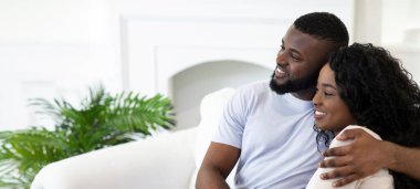 Siyah bir adam ve kadın evlerinde beyaz bir kanepede oturuyorlar. Adamın kolu kadının omzunda ve ikisi de mesafeye bakıyor..