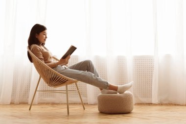 Bir kadın pencere kenarında bir sandalyede oturup kitap okuyor. Ayakları dinleniyor..