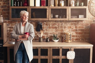 Son sınıf öğrencisi bir kadın, elinde kırmızı bir fincan kahveyle tuğla duvarlarla kırsal bir mutfakta duruyor. Pencereden bakarken gülümsüyor, sabah içkisinin tadını çıkarıyor, boşluğu kopyalıyor.