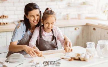 Bir anne ve kızı mutfakta birlikte çalışıyor, kurabiye için hamur topluyor. Anne kolunu kızın omzuna atmış ve eline oklavayı takarken ona rehberlik ediyor.