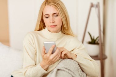 Sarı saçlı bir kadın kanepede oturmuş telefonuna bakıyor..