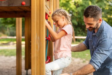 Bir baba, ahşap bir oyun bahçesinde ip merdivene tırmanan kızını cesaretlendirir. Gülümsüyor ve merdivenleri çıkarken onu izliyor..