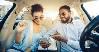 Genç siyah bir çift, güneş gözlüğü ve günlük giysiler takıyor, bir arabanın arka koltuğunda oturuyor ve akıllı telefona bakıyorlar. Gülümsüyorlar ve birbirlerine eşlik etmekten zevk alıyor gibi görünüyorlar.