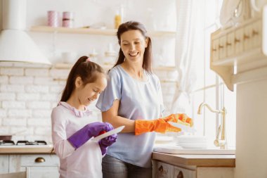 Bu resim modern bir mutfakta bulaşıkları yıkayan bir anne ve kızı gösteriyor. Annem turuncu eldiven giyiyor ve lavaboda bir tabak yıkıyor. Kızı mor eldiven giyiyor ve elinde bir tabak tutuyor.