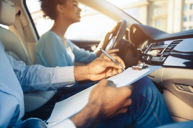 A driving instructor sits in the passenger seat of a car, holding a clipboard and pen, evaluating a student who is behind the wheel. Instructor appears to be taking notes on the students performance clipart