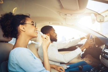 Kıvırcık saçlı genç siyah bir kadın bir arabanın yolcu koltuğunda otururken dudak kremi sürüyor. Arabayı bir adam kullanıyor. Kadın gülümsüyor ve dikiz aynasında yansımasına bakıyor..