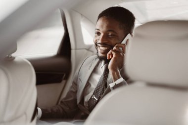 Başarılı bir Afrikalı Amerikalı iş adamı, takım elbise ve kravat takıyor, arabanın arka koltuğunda oturuyor ve gülümserken telefonla konuşuyor..