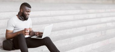 Beyaz tişörtlü bir adam basamaklarda oturuyor, dizüstü bilgisayar kullanıyor ve elinde bir fincan kahve tutuyor..