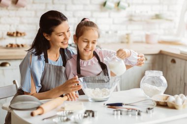 Bir anne ve küçük kızı mutfakta birlikte yemek pişiriyorlar. Annesi gülümseyerek izlerken kız bir kase una süt döküyor..