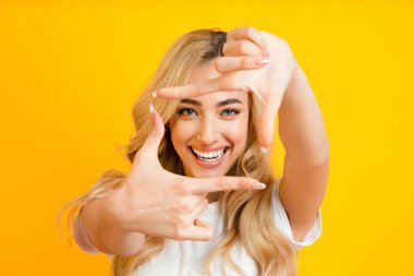 Parlak bir gülüşü olan genç sarışın bir kadın sarı arka planda duruyor ve elleriyle bir çerçeve yapıyor. Beyaz bir tişört giyiyor ve saçları omuzlarından süzülüyor..