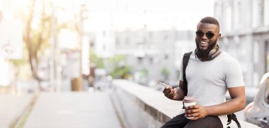 Güneş gözlüğü takan ve sırt çantası takan bir adam şehir ortamında bir çıkıntıda oturur, elinde kahve fincanı ve akıllı telefonuyla..