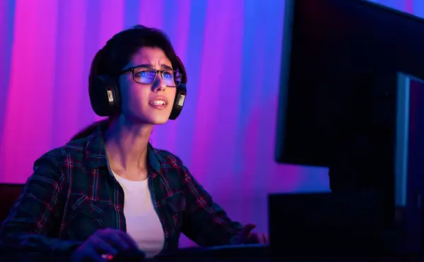 stock image A young woman with glasses and headphones is seated in front of a computer screen. She appears to be focused intently on the screen, with a serious expression on her face