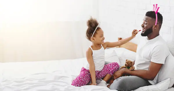 stock image African American little girl, wearing a white tank top and pink floral pants, is applying lipstick to her fathers lips. They are both sitting on a white bed, and the girl is smiling.