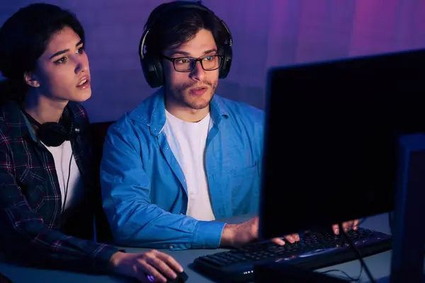 stock image A young man and woman are sitting at a desk in front of a desktop computer, both wearing headphones and looking intently at the screen.