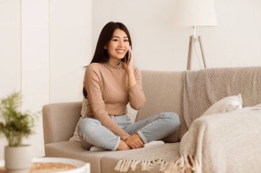 Oturma odasındaki kanepede oturan genç bir kadın cep telefonuyla konuşuyor ve gülümsüyor..