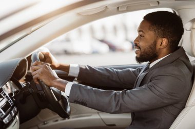 Takım elbiseli profesyonel bir Afro-Amerikan adam odaklanmış ve kendinden emin bir ifadeye sahip bir araba kullanıyor. Şoför koltuğunda oturuyor, sağ eliyle direksiyonu tutuyor.
