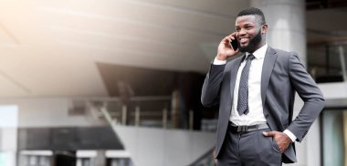 Çekici Afrikalı genç iş adamı telefon ediyor. Ofis binasının yanında eli cebinde duruyor.