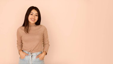 Şeftali rengi bir arka planın önünde duran uzun siyah saçlı genç bir kadın gülümsüyor. Uzun kollu kahverengi bir kazak ve elleri cebinde açık mavi kot pantolon giyiyor..