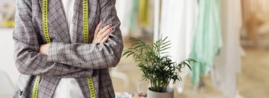 Bir kadın bir tasarım stüdyosunda dikiliyor, boynunda sarı bir mezura olan ekose bir ceket giyiyor..