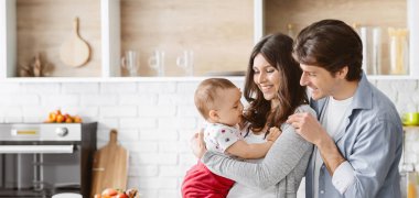 Bu resim, üç kişilik mutlu bir aileyi mutfaklarında gösteriyor. Ebeveynler gülümsüyor ve bebeklerini kollarında tutuyorlar. Bebek ebeveynlerine bakıyor ve mutlu görünüyor..