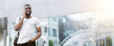 Beyaz tişörtlü bir adam telefonuyla konuşurken şehir caddesinde yürüyor. Gülümsüyor ve yürürken yukarı bakıyor..