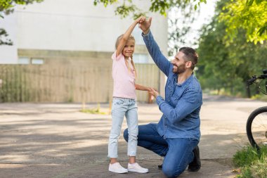 Bir baba güneşli bir günde kızıyla oynamak için diz çöker. İkisi de gülümsüyor ve dışarıda eğleniyorlar. Arka planda bisiklet var..